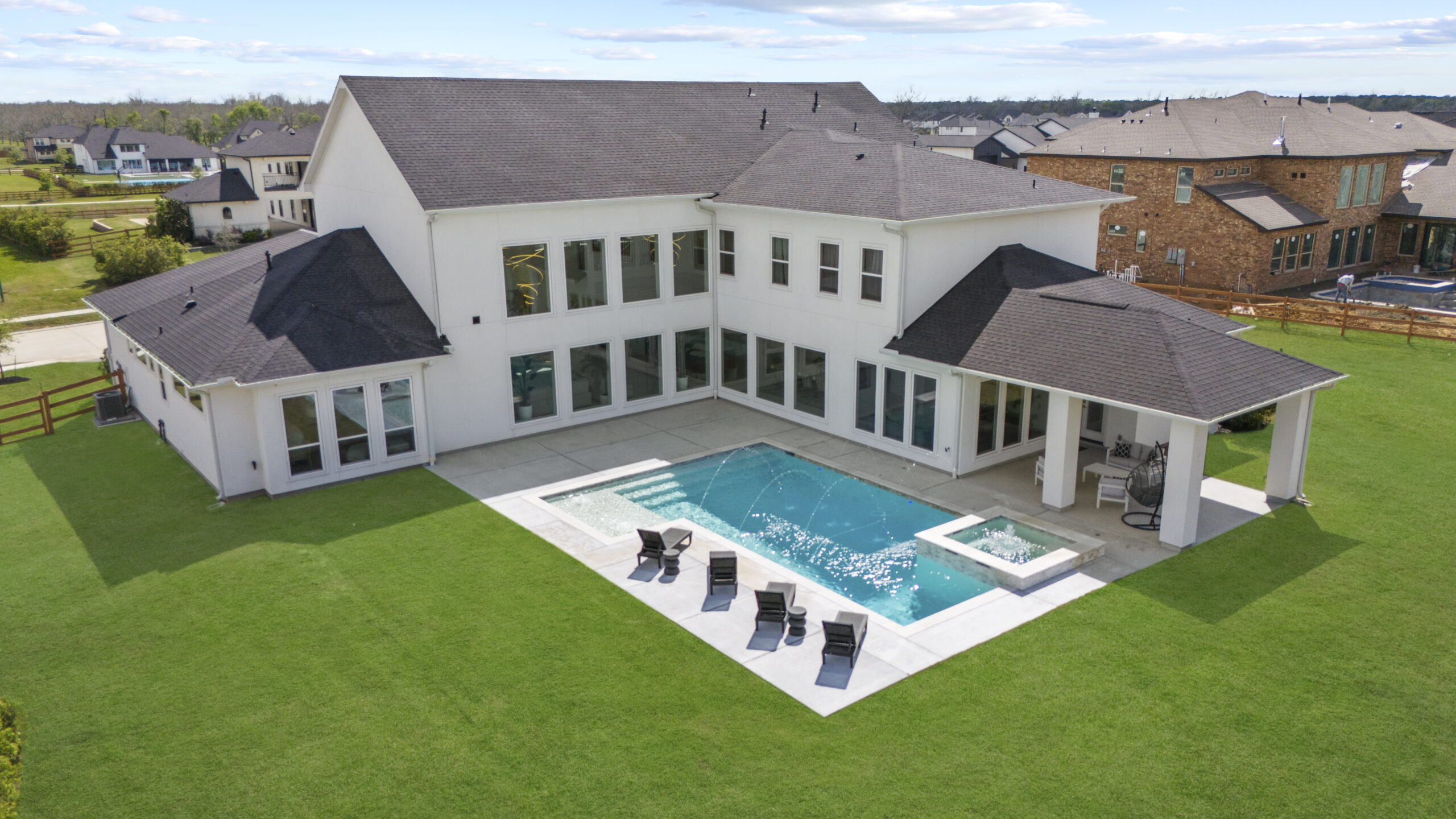 Aerial view of a luxury Houston home for sale with a white stucco exterior, geometric pool with deck jets, spa, covered patio, and large landscaped lawn.