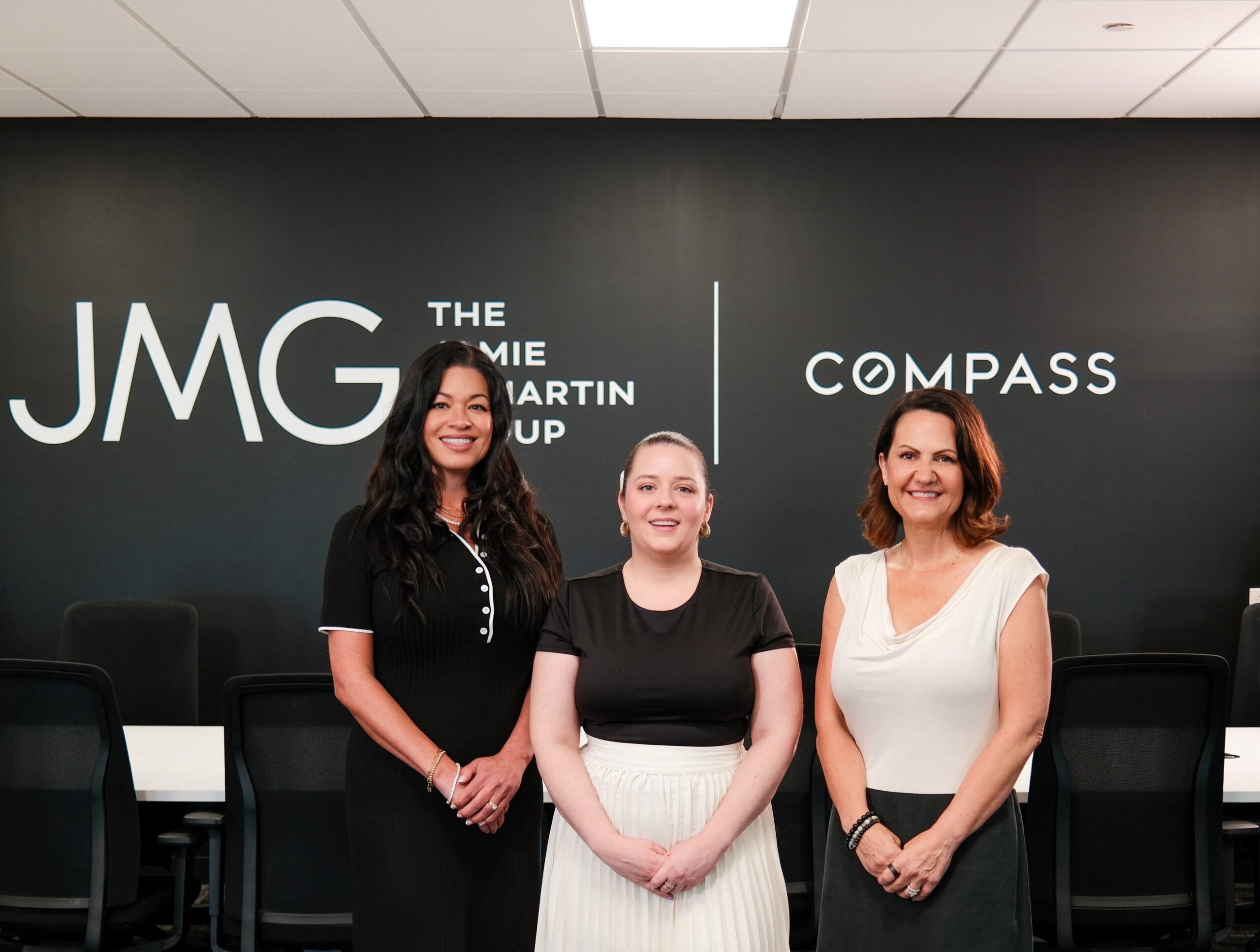 Three members of The Jamie McMartin Group standing in front of the JMG and Compass logo wall at the team's Houston-area office.