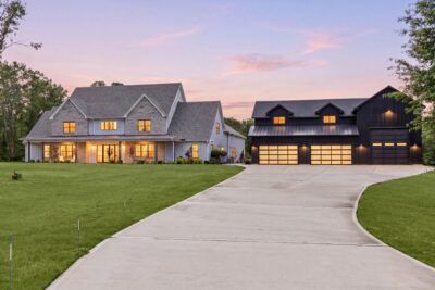 A full-width covered porch, and a standing-seam metal porch roof anchor the main residence, with a separate garage and workshop building set just across the drive and wooded green space beyond.