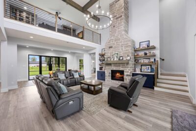 The living area rises a full two stories, centered on a floor-to-ceiling stone gas log fireplace with a wood mantel, flanking built-ins, and floating shelves. Exposed beams and a loft overlook frame the space from above.