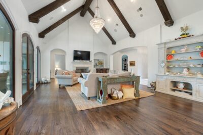 Living area with vaulted ceilings, exposed wood beams, stone fireplace, crystal chandelier, and refinished hardwood floors creating a warm, elegant space.