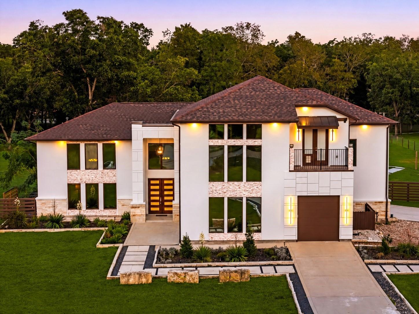 Contemporary home for sale in Fulshear at twilight, with a white stucco and stone facade, double wood front doors, a second-floor balcony and a landscaped front yard backed by mature trees