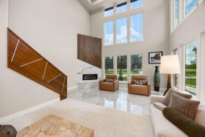 Two-story living room in a home for sale in Fulshear, with a wall of windows, marble floors, a linear fireplace with marble surround and slatted wood accent, and a wood-paneled staircase
