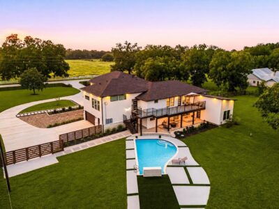 Twilight aerial view of a home for sale in Fulshear, showing the pool with stone water feature, covered patio, expansive second-story deck and fenced backyard with open pasture beyond
